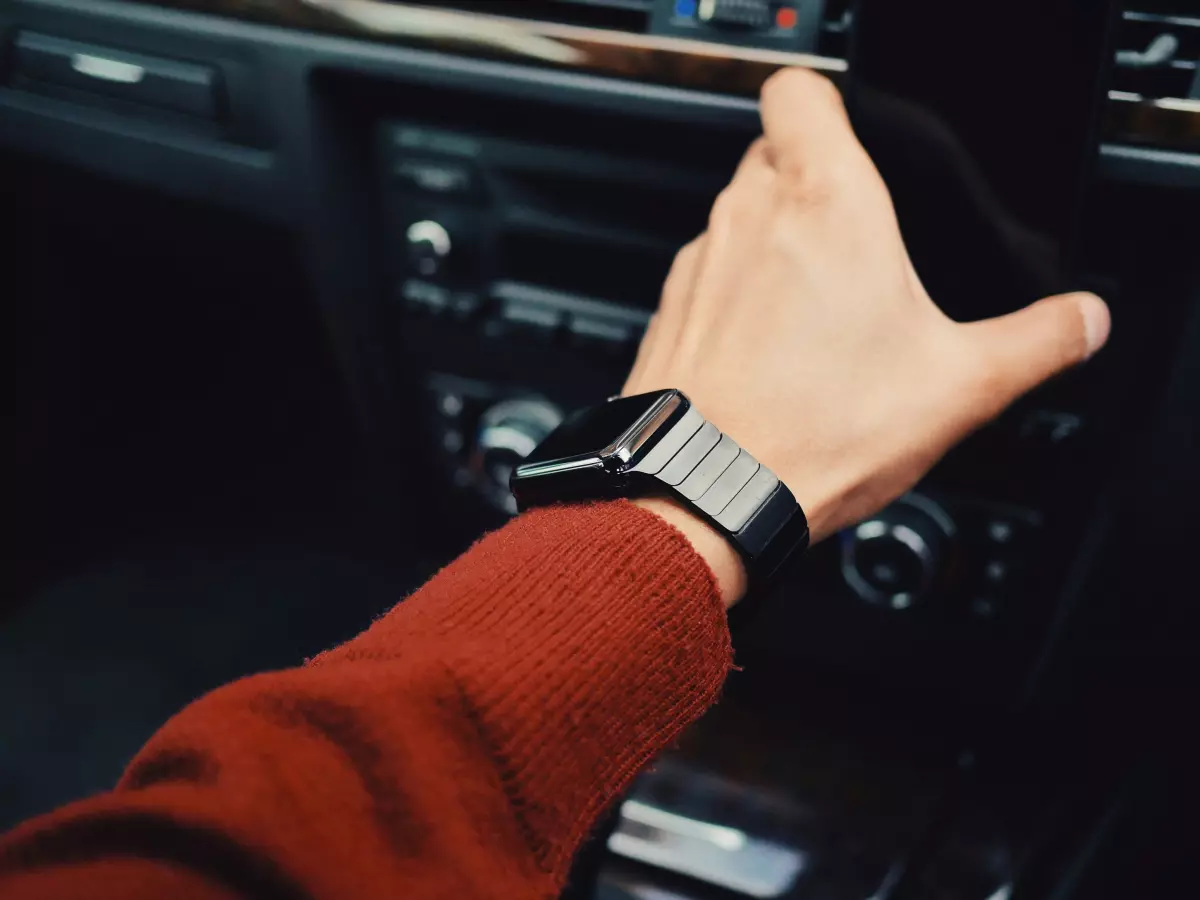 A person's hand wearing a smartwatch, with a red shirt, is reaching to adjust a car radio, suggesting the smartwatch's compatibility and usefulness.