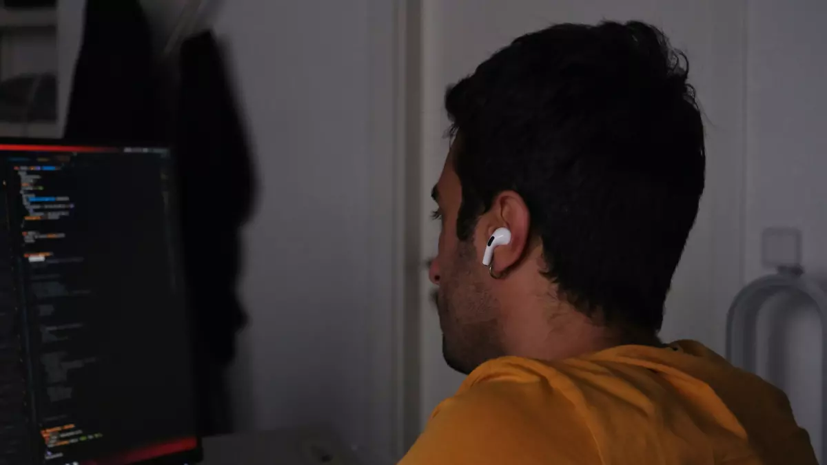 A young man with dark hair is sitting in front of a computer, wearing a yellow shirt. He is typing on the keyboard. His face is not visible, but he seems to be concentrating on his work.