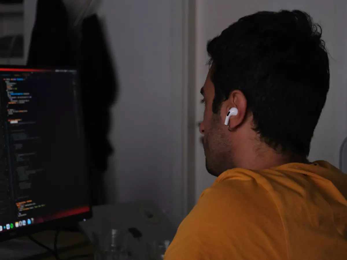 A young man with dark hair is sitting in front of a computer, wearing a yellow shirt. He is typing on the keyboard. His face is not visible, but he seems to be concentrating on his work.