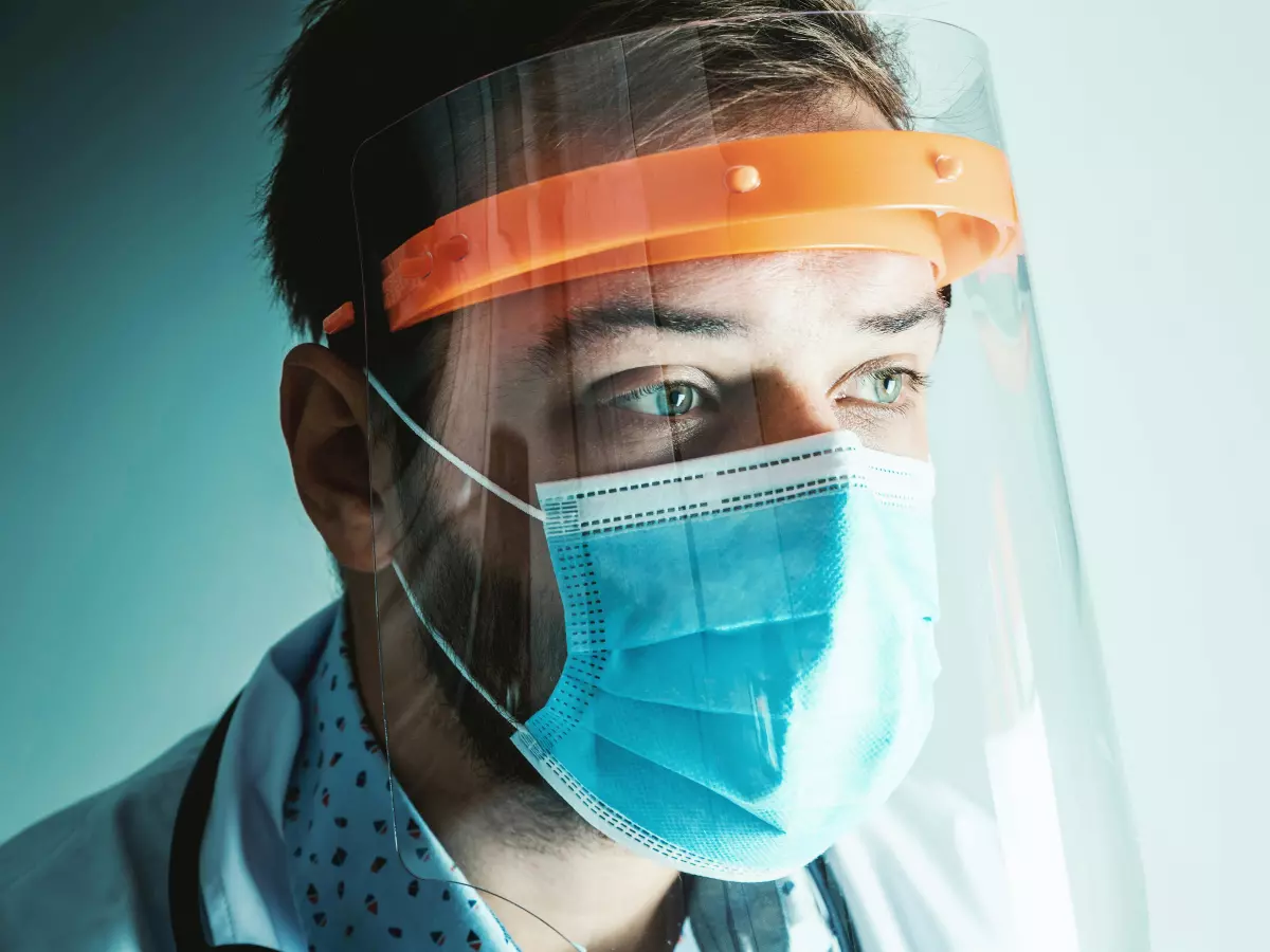 A doctor wearing a face shield and a mask looks thoughtfully ahead.