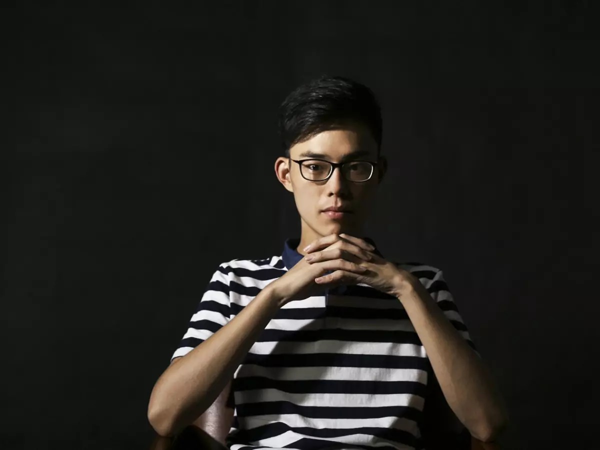 A young man wearing glasses is sitting in a chair. He is wearing a striped shirt and his arms are crossed on his lap. The background is black.