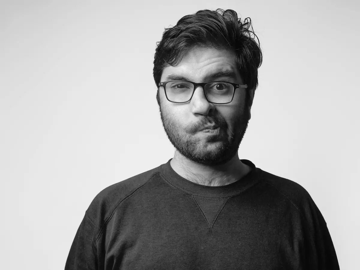 A man with a beard and glasses is looking at the camera with a confused expression, his lips slightly pursed. He is wearing a black sweater and has dark hair. The background is white.