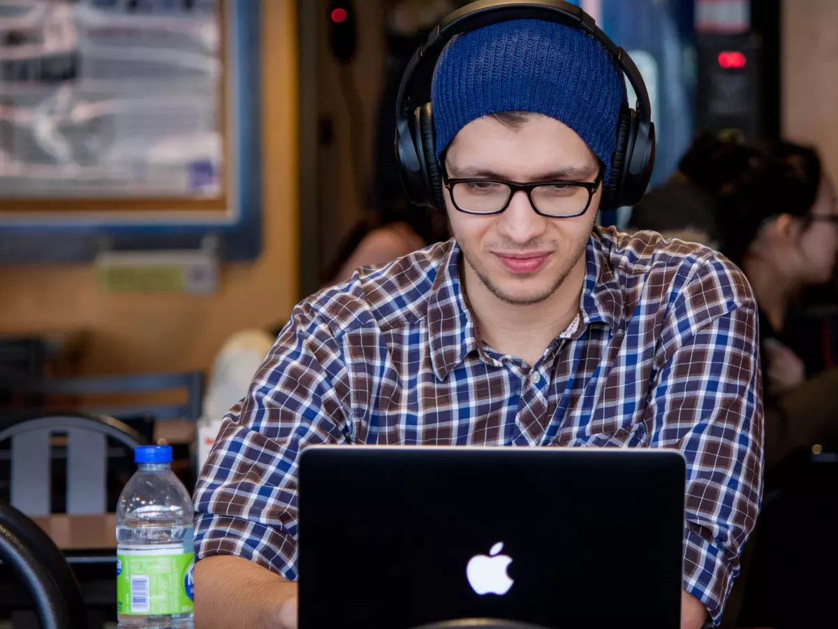 A young man in a blue beanie and headphones is sitting in a coffee shop and working on a laptop.