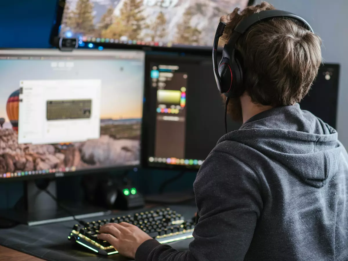 A person wearing headphones is looking at a computer screen with multiple monitors and using a keyboard and mouse. The screen shows a game interface.