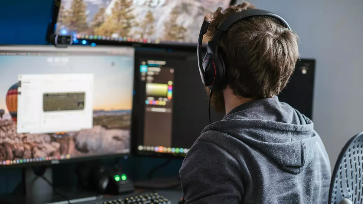 A person wearing headphones is looking at a computer screen with multiple monitors and using a keyboard and mouse. The screen shows a game interface.