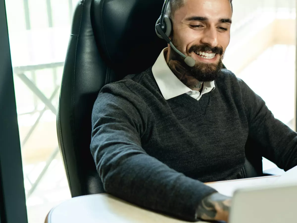 A young man with a beard, wearing a black sweater, headset, and a tattoo on his hand, looking at a laptop in a modern office.