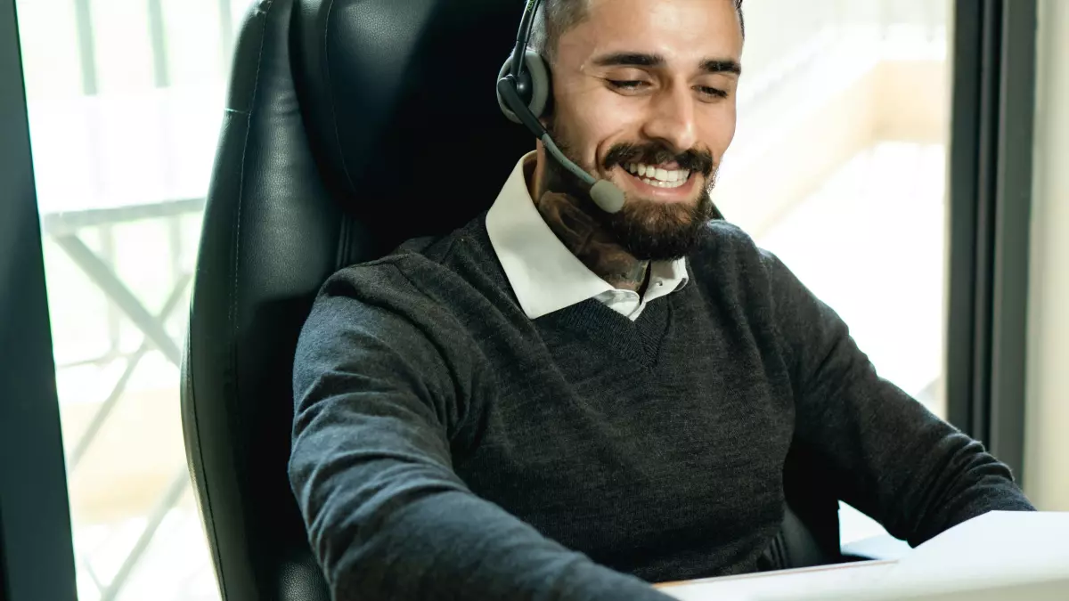 A young man with a beard, wearing a black sweater, headset, and a tattoo on his hand, looking at a laptop in a modern office.