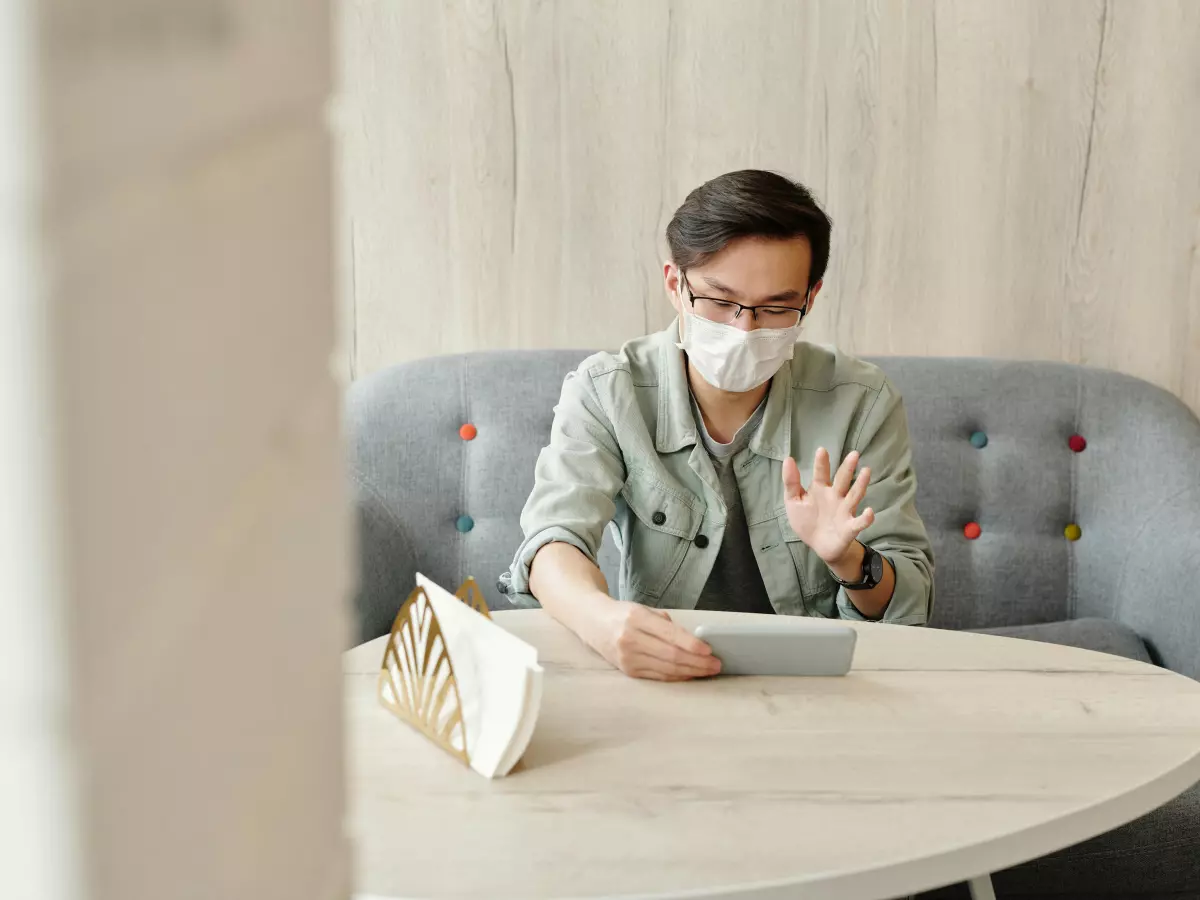 A person wearing a face mask and glasses is sitting on a couch and using a tablet to video call someone. 