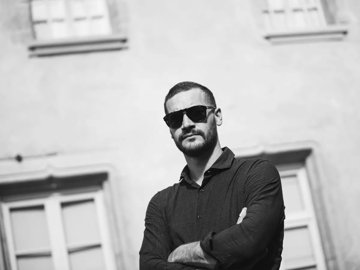 A man with a beard wearing sunglasses stands with his arms crossed in front of a building.