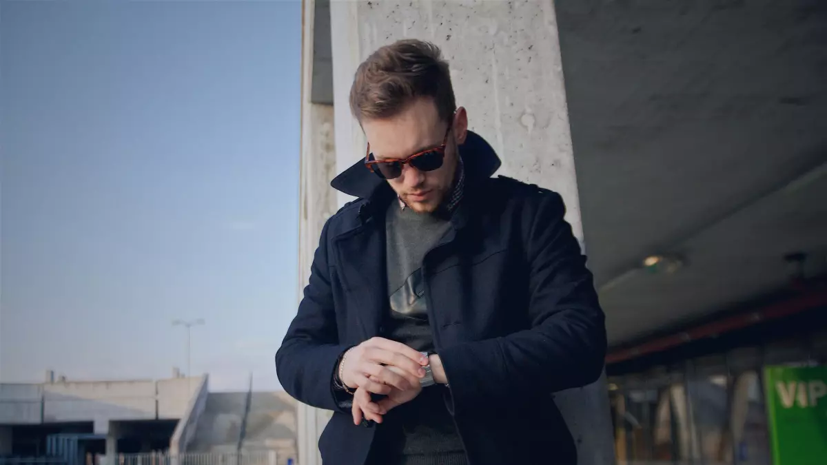 A man wearing a blue coat and sunglasses looks at his watch.