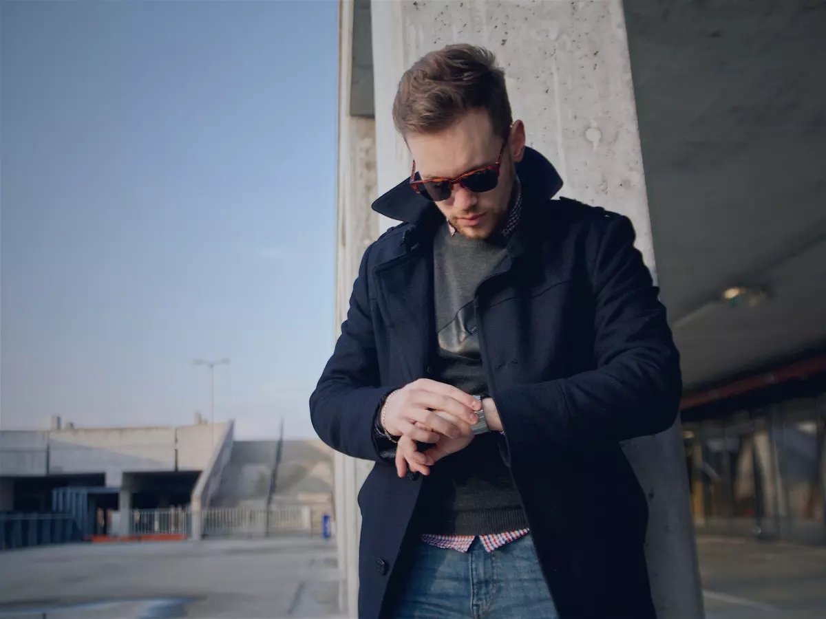 A man wearing a blue coat and sunglasses looks at his watch.