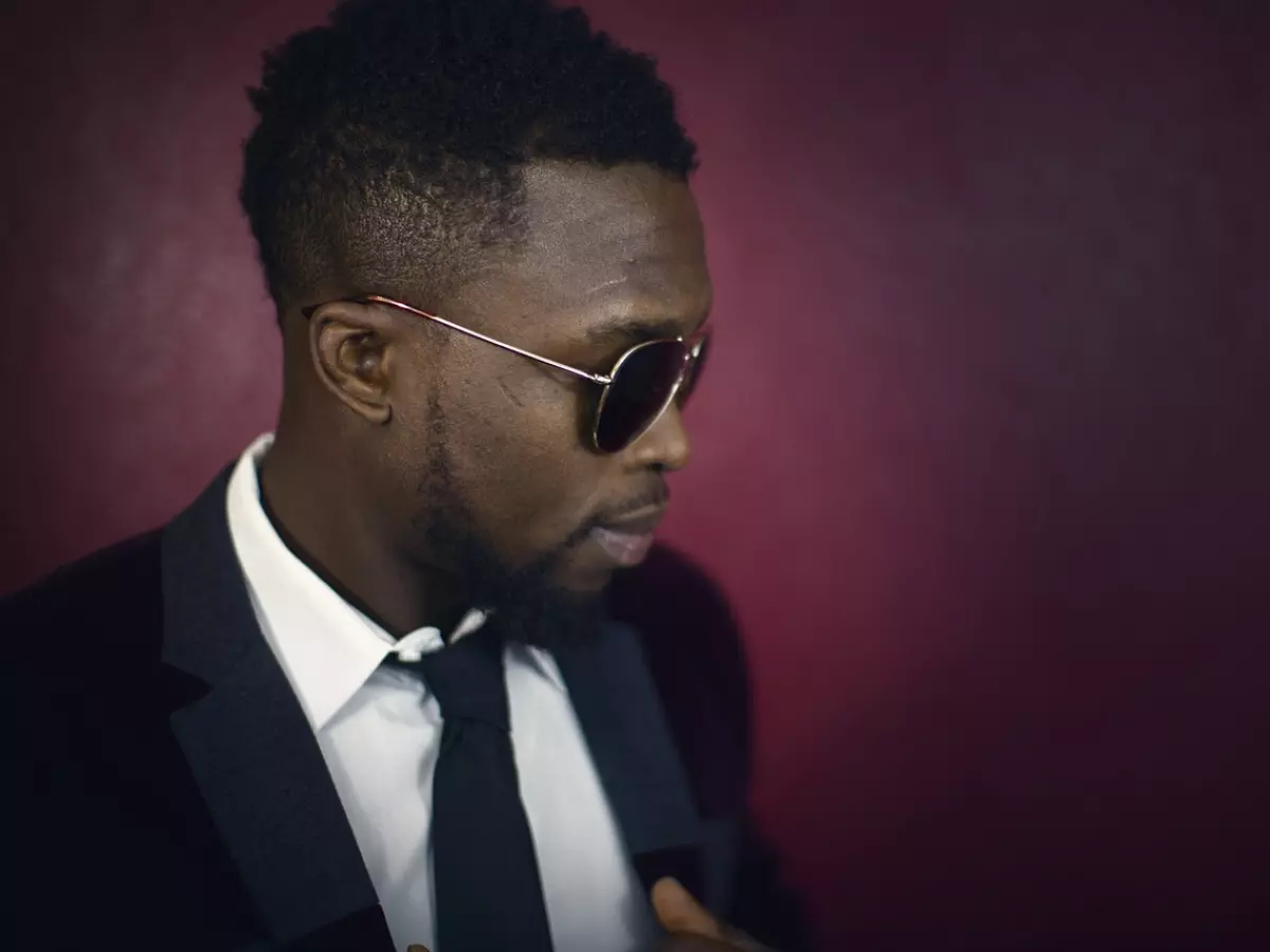 A man wearing a dark suit and sunglasses is looking to the side. He is standing in front of a dark red wall.