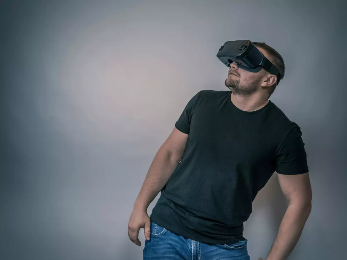 A man wearing VR glasses stands in front of a grey wall, looking up.