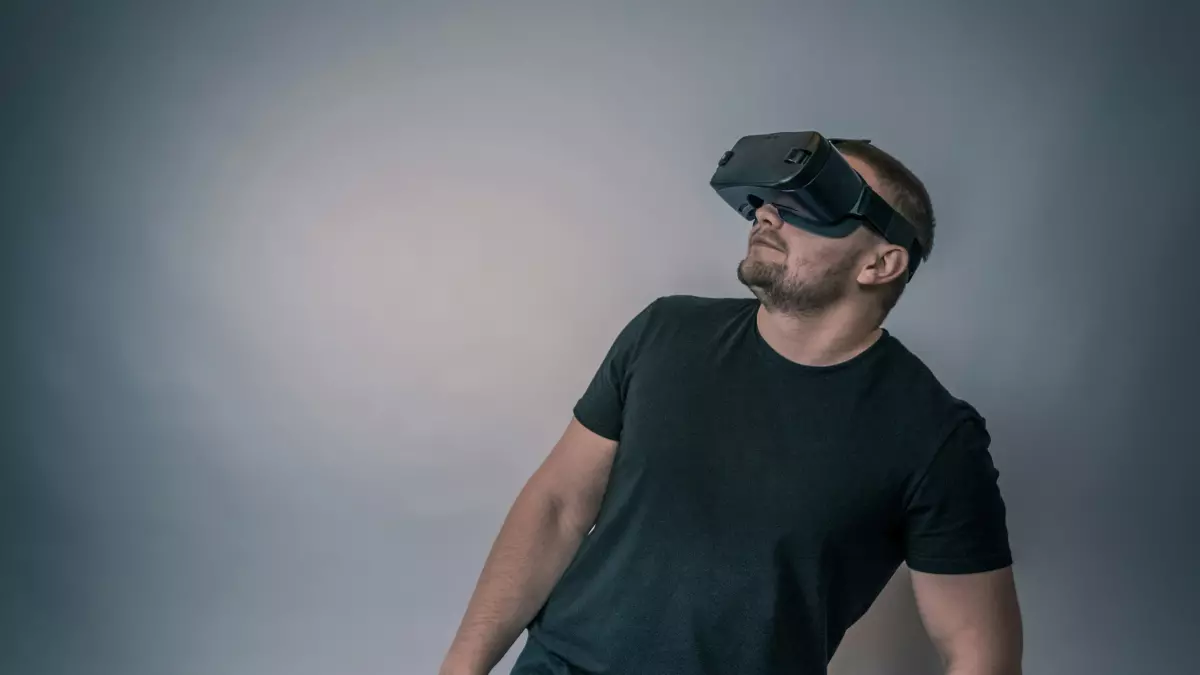 A man wearing VR glasses stands in front of a grey wall, looking up.