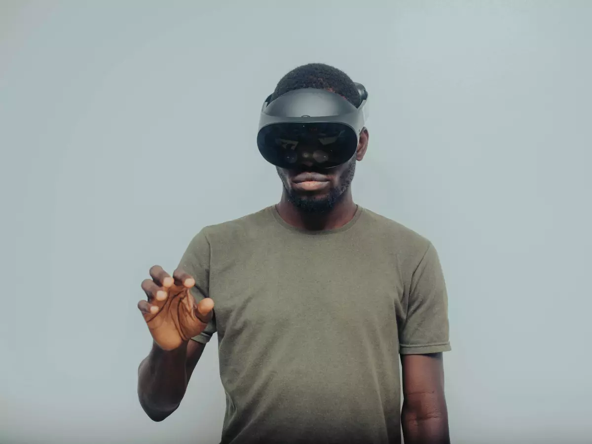 A young African American man with a beard wearing a VR headset. He's looking straight into the camera and his hand is outstretched as if reaching for something. The background is a plain white wall.