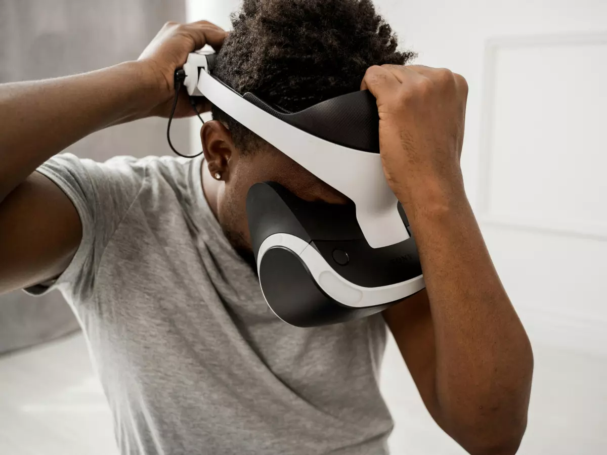 A man is wearing a virtual reality headset. He is holding the headset with both hands.  The room is white with a window in the background.