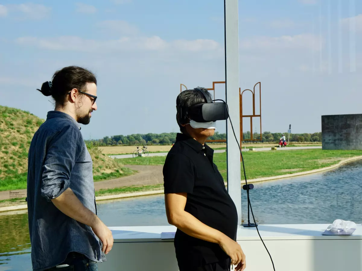 A man wearing a VR headset experiences the virtual world, while another man stands nearby watching.