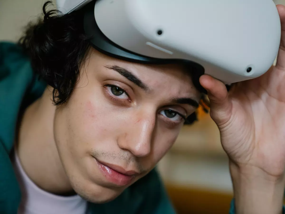 A close-up of a young man wearing a white VR headset. He is looking directly at the camera with a neutral expression. His hand is touching the headset.