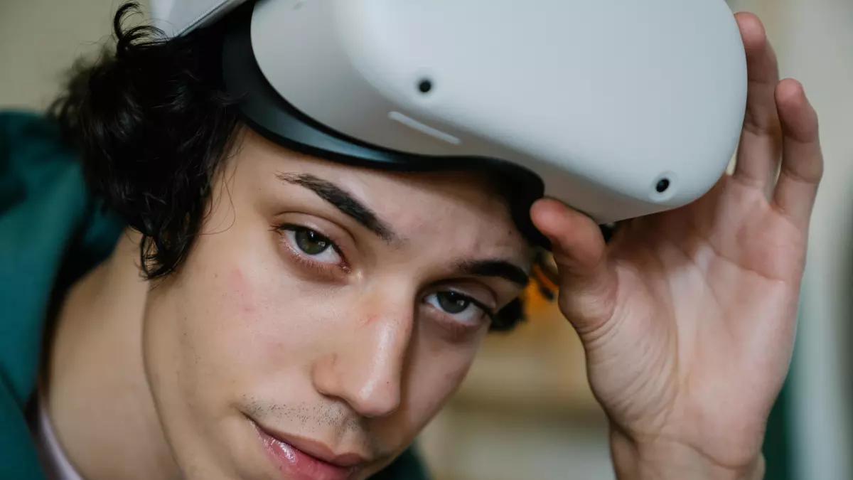 A close-up of a young man wearing a white VR headset. He is looking directly at the camera with a neutral expression. His hand is touching the headset.