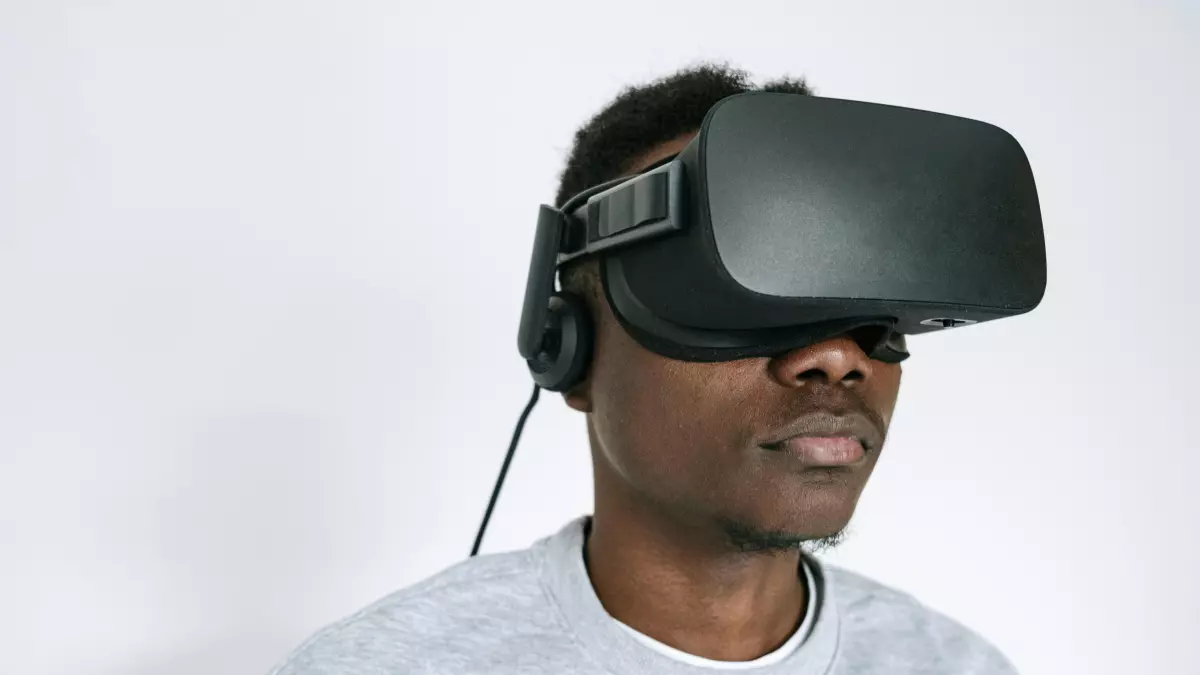 A man is wearing a black VR headset and looking straight ahead. He is wearing a grey sweater and the background is a white wall.