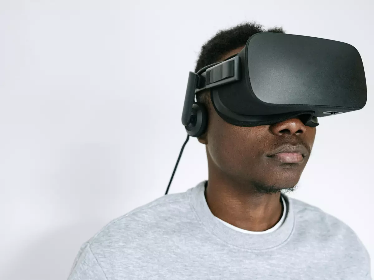 A man is wearing a black VR headset and looking straight ahead. He is wearing a grey sweater and the background is a white wall.