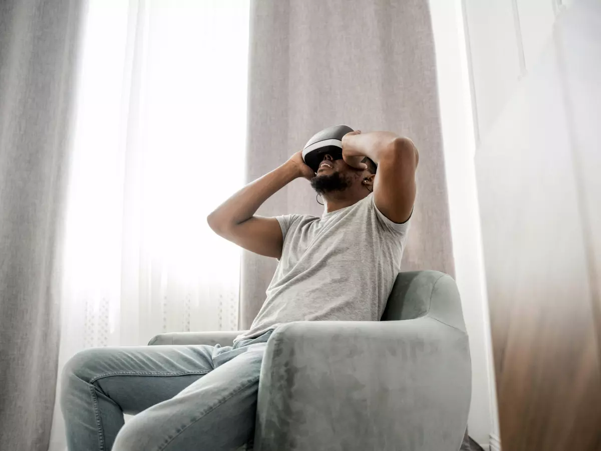 A man in a grey t-shirt and jeans is sitting in an armchair. He has his hands on his head and appears to be thinking or relaxing. He's sitting in front of a window with white curtains.