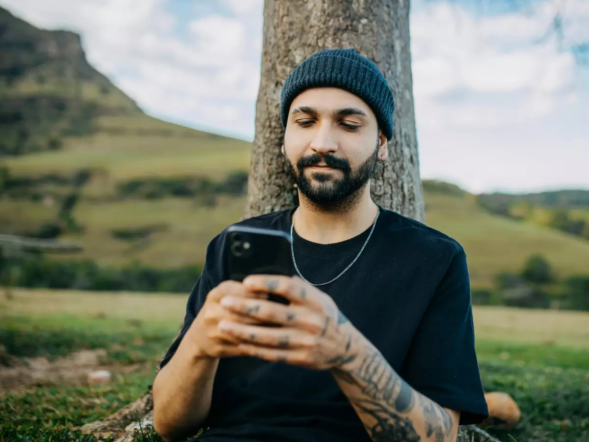 A man wearing a black beanie and black t-shirt with tattoos on his arms is leaning against a tree and looking down at his phone.