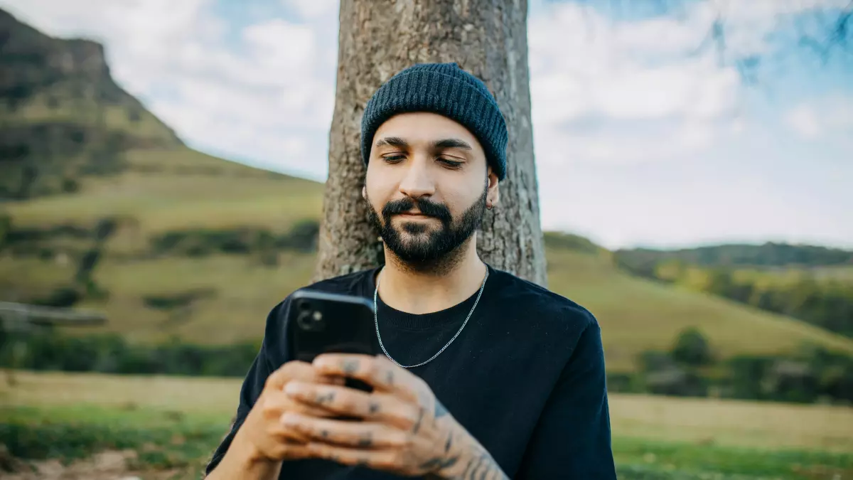 A man wearing a black beanie and black t-shirt with tattoos on his arms is leaning against a tree and looking down at his phone.