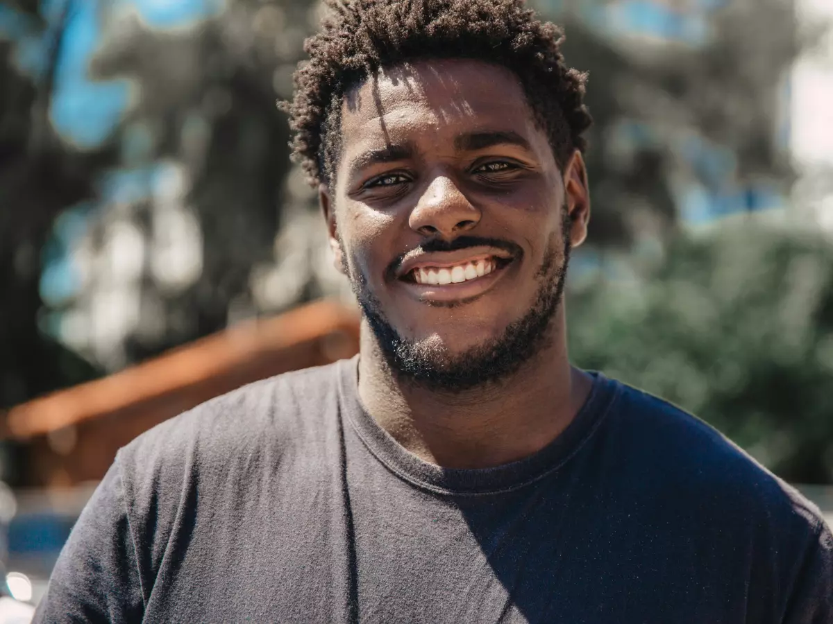 A young black man with short, curly hair and a beard is smiling broadly at the camera. He is wearing a dark blue t-shirt and standing outdoors.