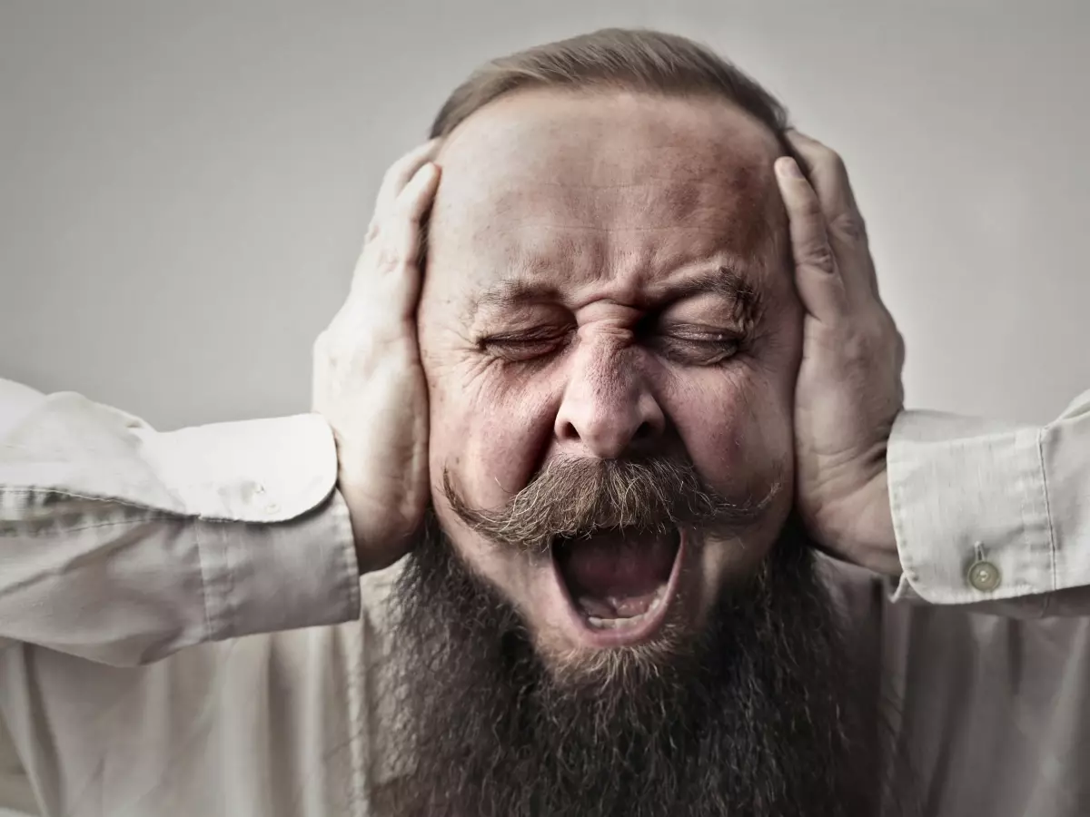 A man with a beard and a white shirt screams and covers his ears with his hands, appearing distressed and frustrated.