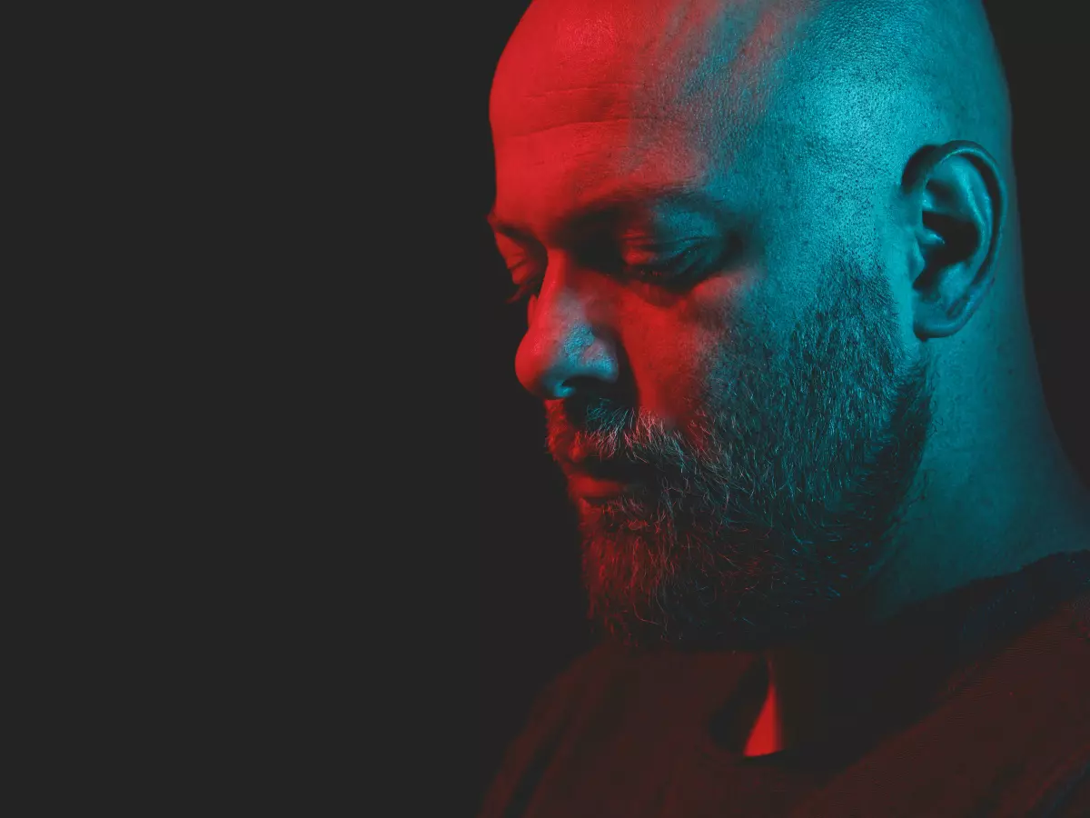 A close-up portrait of a bald man with a beard, illuminated by red and blue lights, looking down.