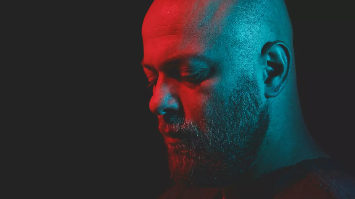 A close-up portrait of a bald man with a beard, illuminated by red and blue lights, looking down.