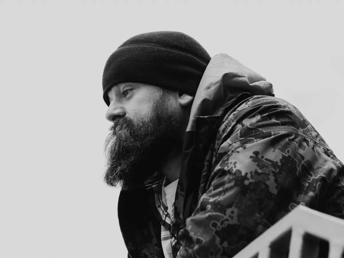 A bearded man in a jacket and beanie looks out into the distance, his expression contemplative and uncertain.