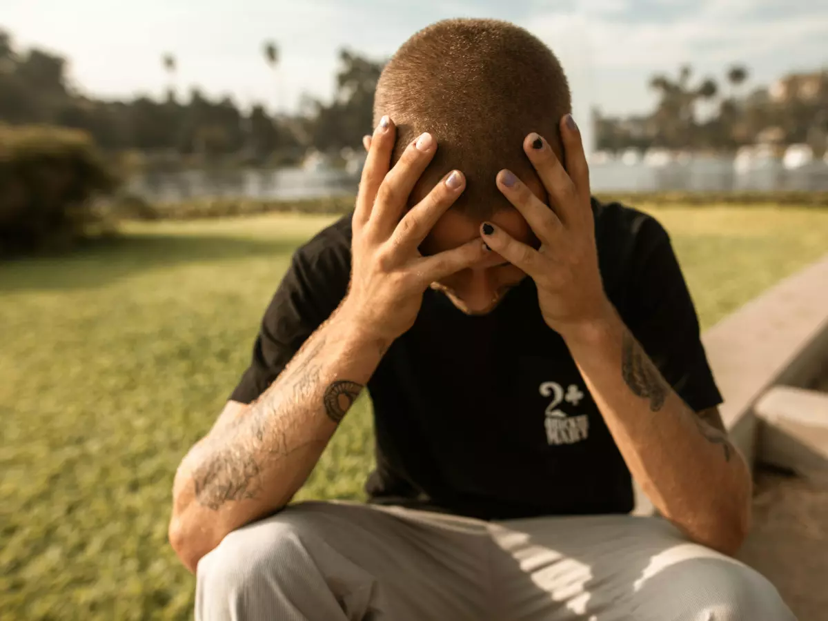 A man sits on a curb with his head in his hands, looking distressed.