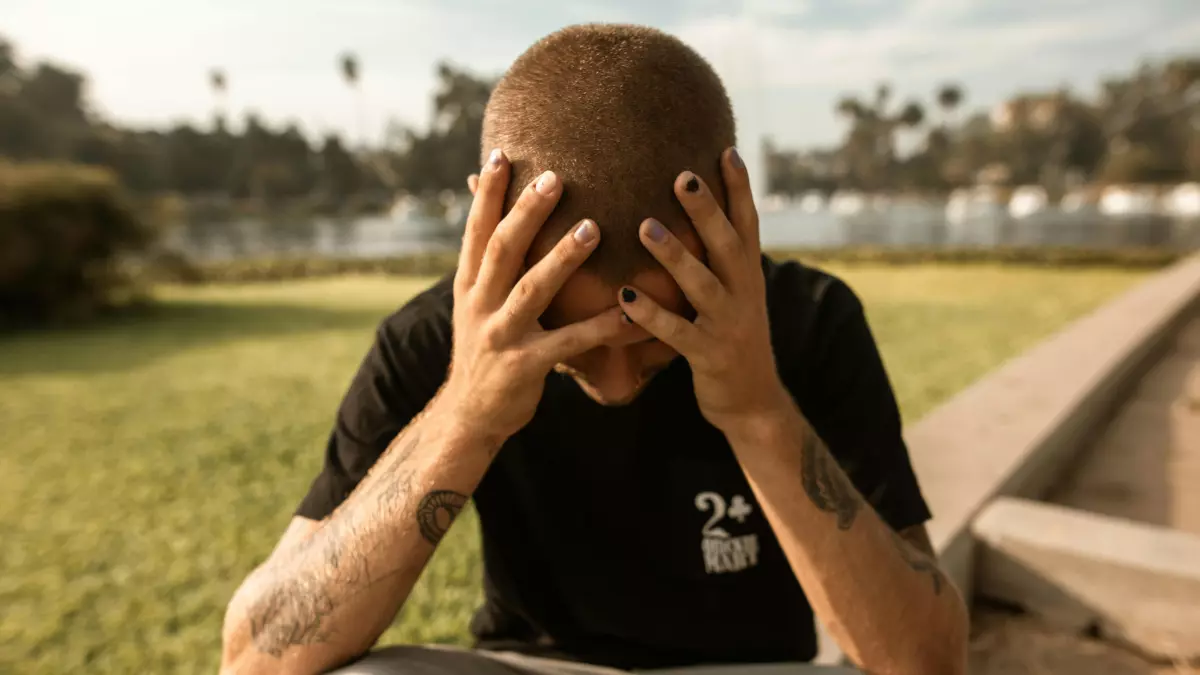 A man sits on a curb with his head in his hands, looking distressed.