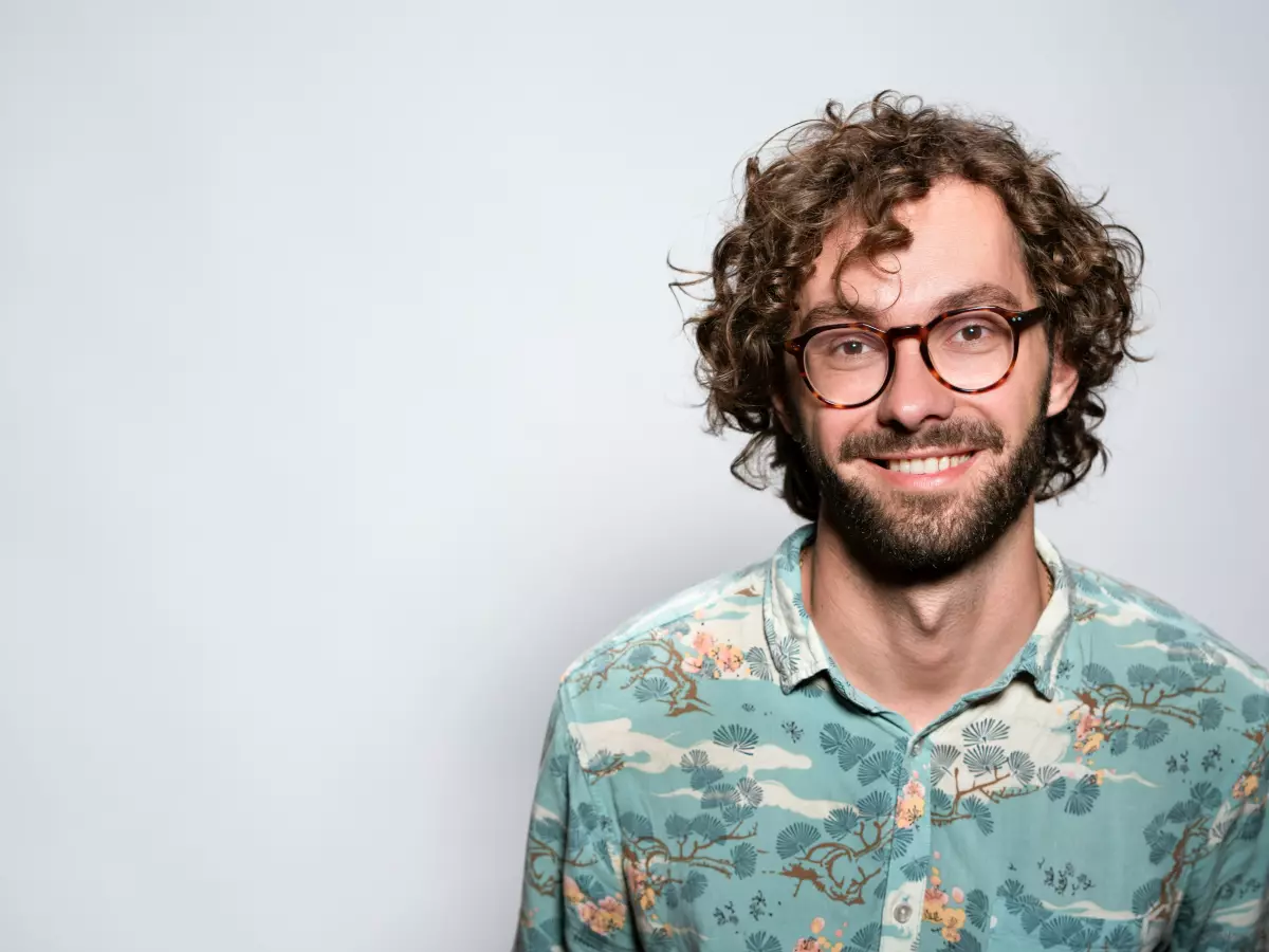 A man with curly hair and a beard is smiling at the camera. He is wearing a blue and white floral shirt and glasses.