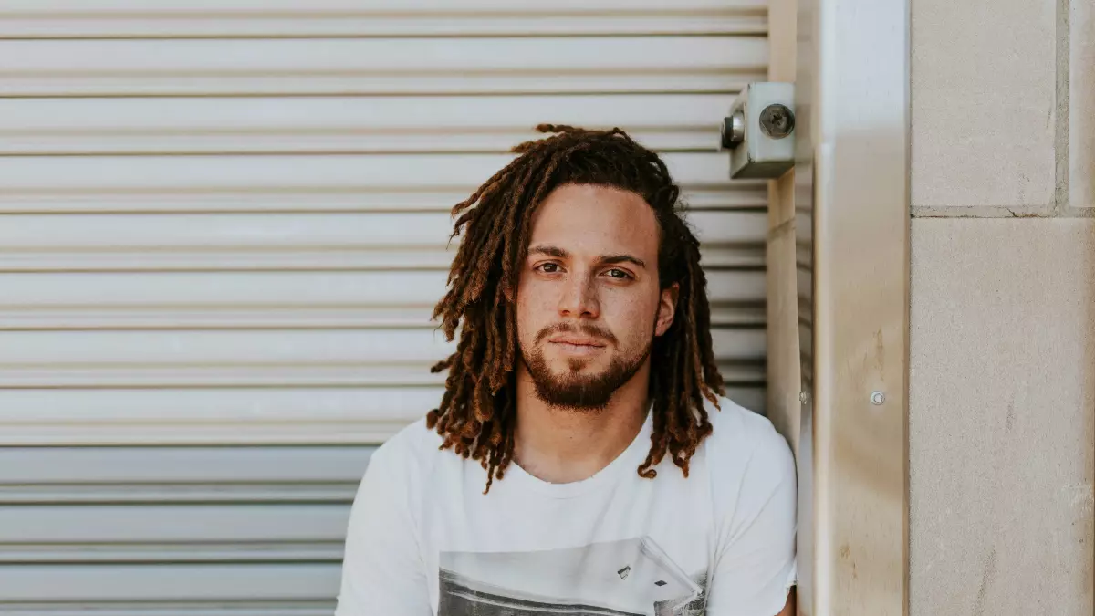 A man with long dreadlocks is sitting in front of a white garage door, wearing a white t-shirt with a printed design.