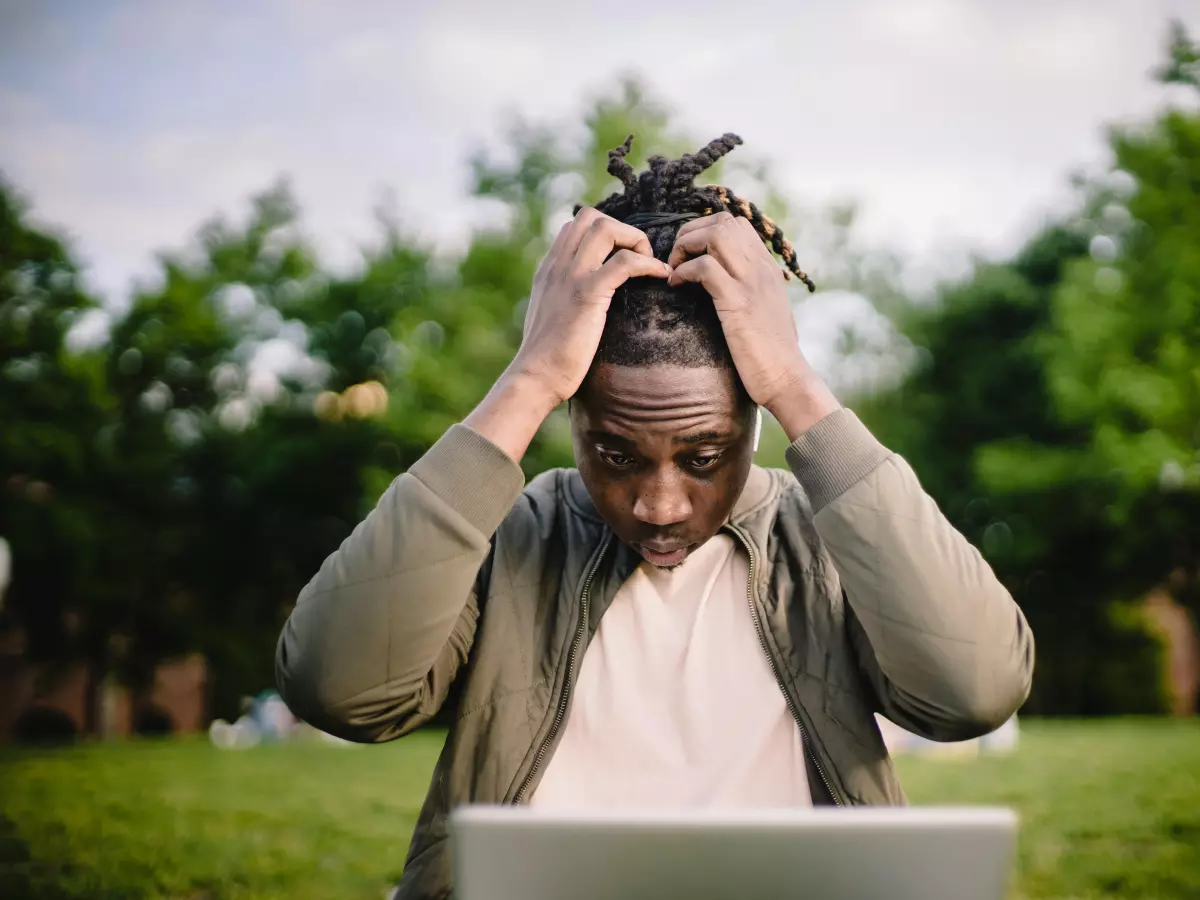 A young man sits in front of a laptop with a frustrated expression, clutching his head with both hands.