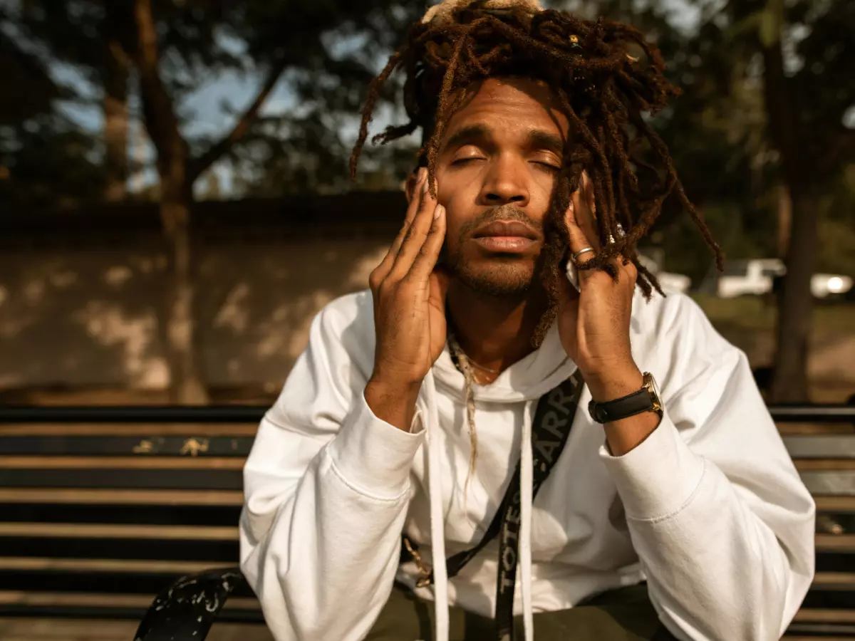 A young man with dreadlocks sits on a park bench with his hands on his face, looking stressed. He is wearing a white hoodie and has a black backpack on.