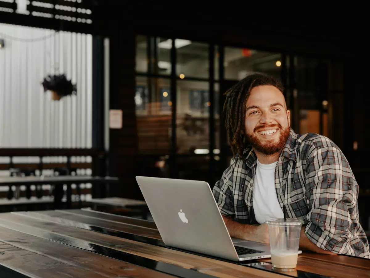 A man in a plaid shirt is sitting at a table with a laptop in front of him. He is smiling and looking to the side.