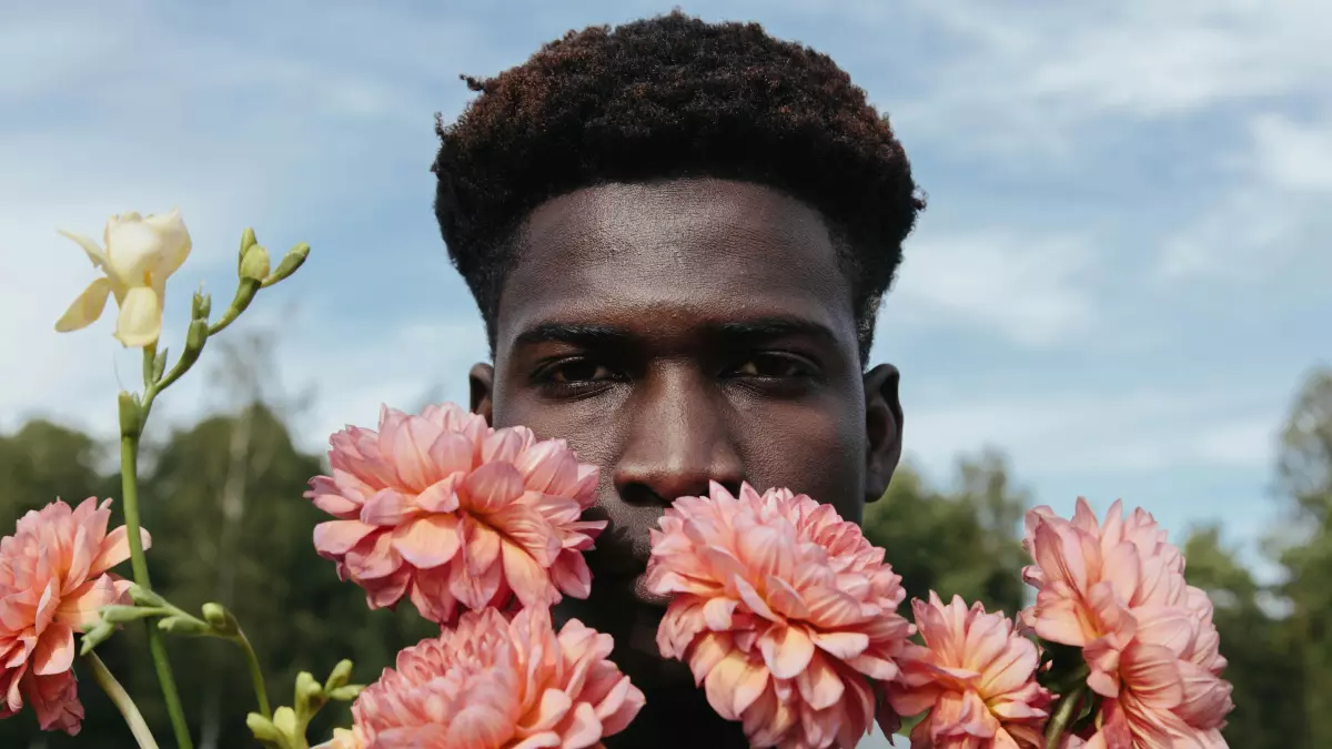 A man with short black hair is standing in a field with pink flowers. He is looking at the camera with a serious expression.