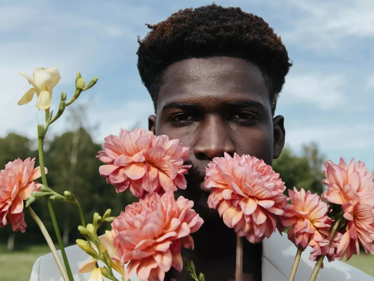 A man with short black hair is standing in a field with pink flowers. He is looking at the camera with a serious expression.