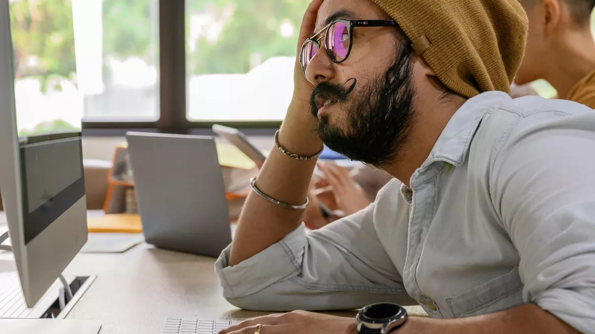 A man with a beard and glasses is sitting at a desk in an office, staring intently at his computer screen. He has a serious expression on his face and his body language is tense. There are other people working in the background, but the man is the focus of the image.