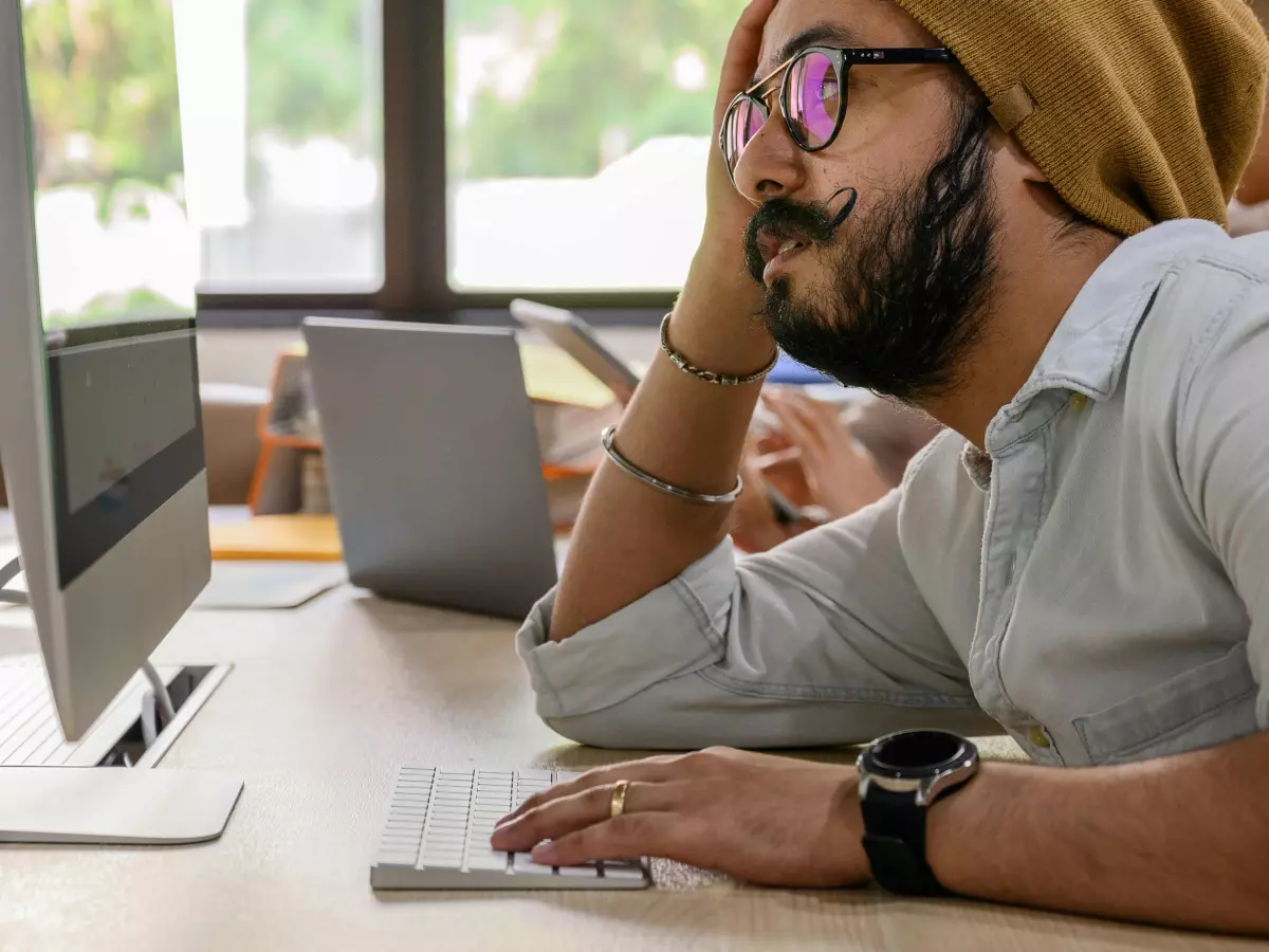 A man with a beard and glasses is sitting at a desk in an office, staring intently at his computer screen. He has a serious expression on his face and his body language is tense. There are other people working in the background, but the man is the focus of the image.