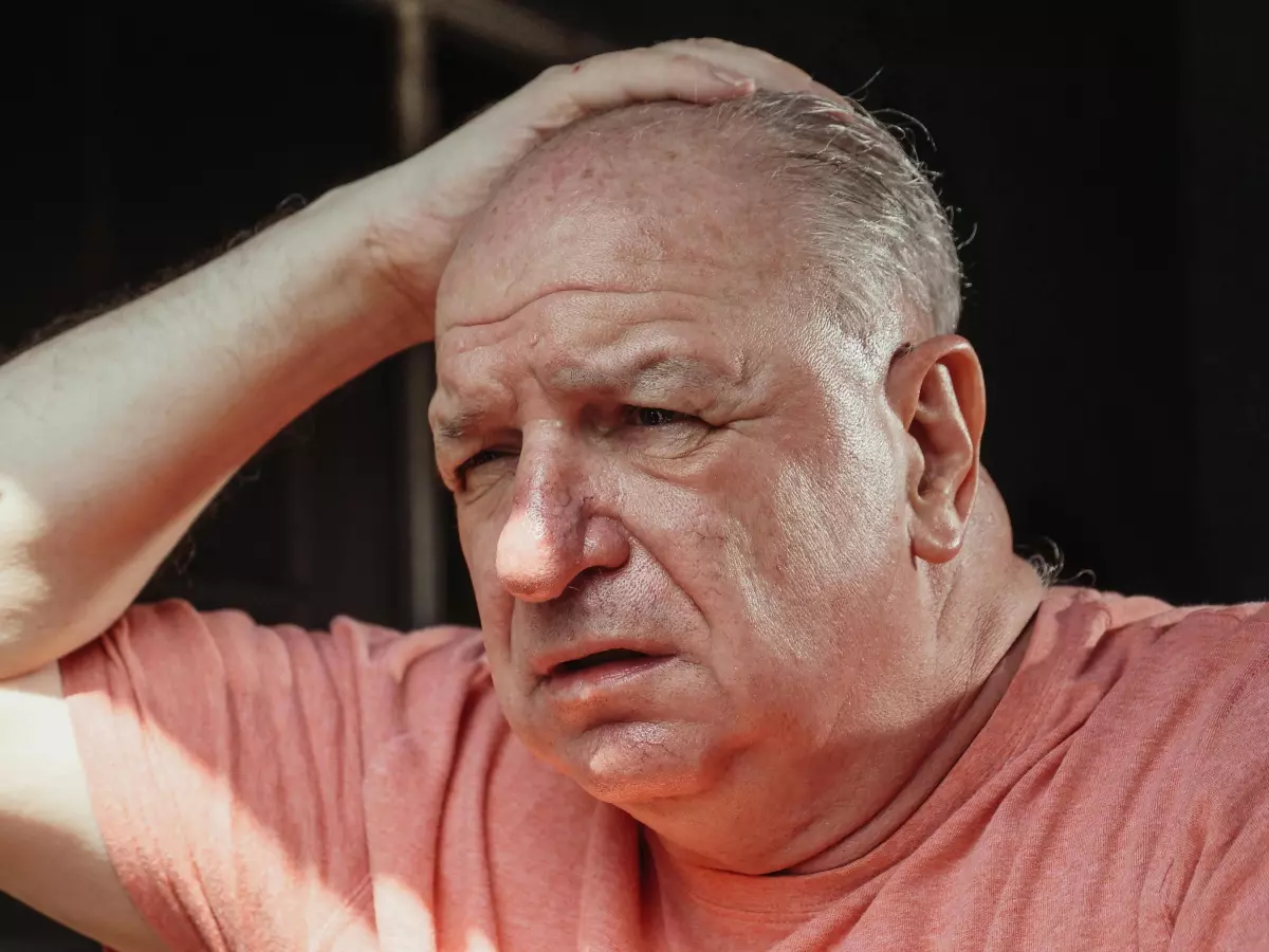 An older man with a pink shirt is looking distraught, grabbing his head with his hand.