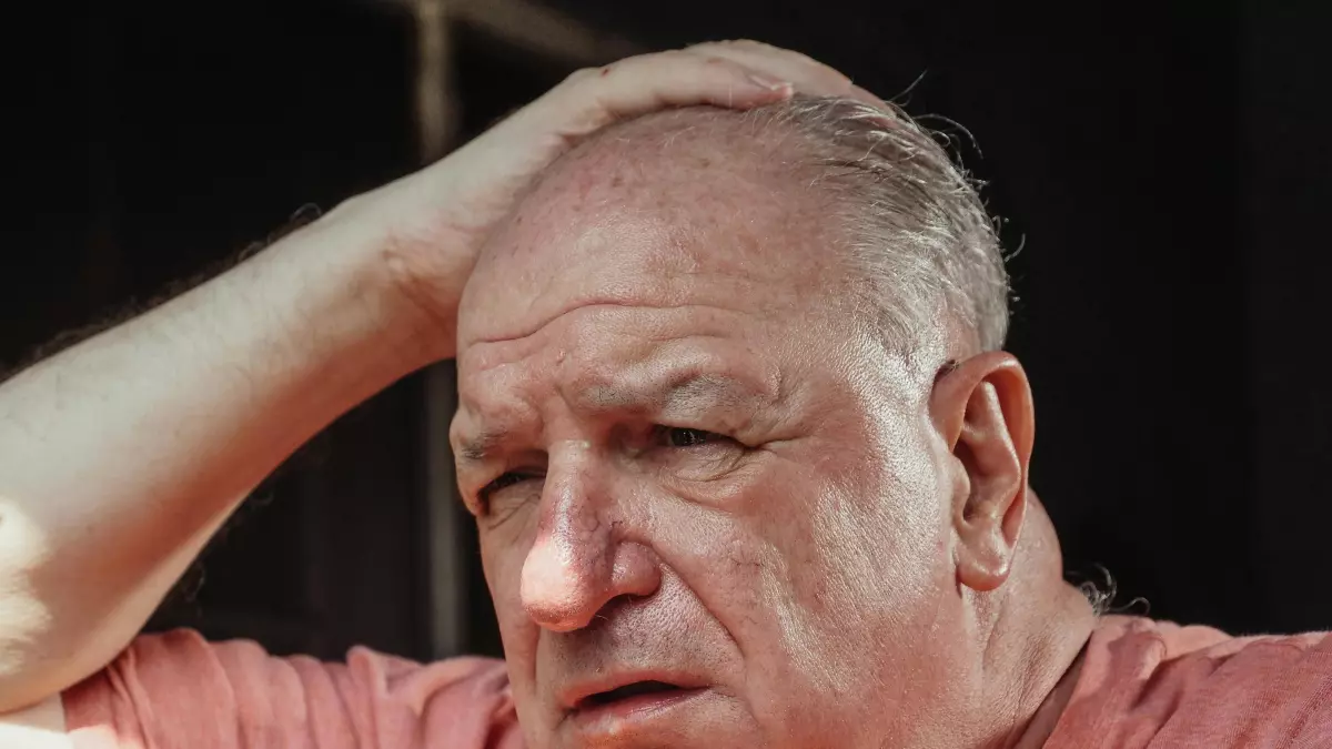 An older man with a pink shirt is looking distraught, grabbing his head with his hand.