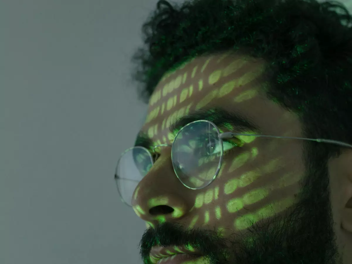 Close-up of a man with a beard wearing glasses.  Green lines resembling code are projected onto his face. He looks intensely at something off-screen.