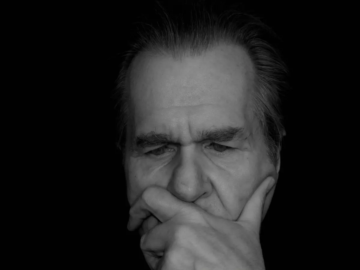 A close-up of an older man's face, in black and white, with his eyes closed and a hand resting on his chin, conveying a sense of worry and contemplation.