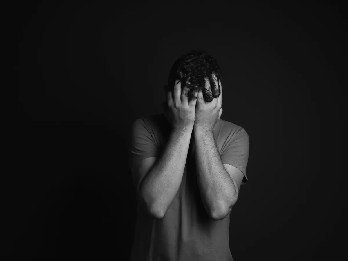 A young man is sitting on a couch, looking down at his phone. He is wearing a blue shirt and jeans. His face is obscured by his hand, and he appears to be in distress.