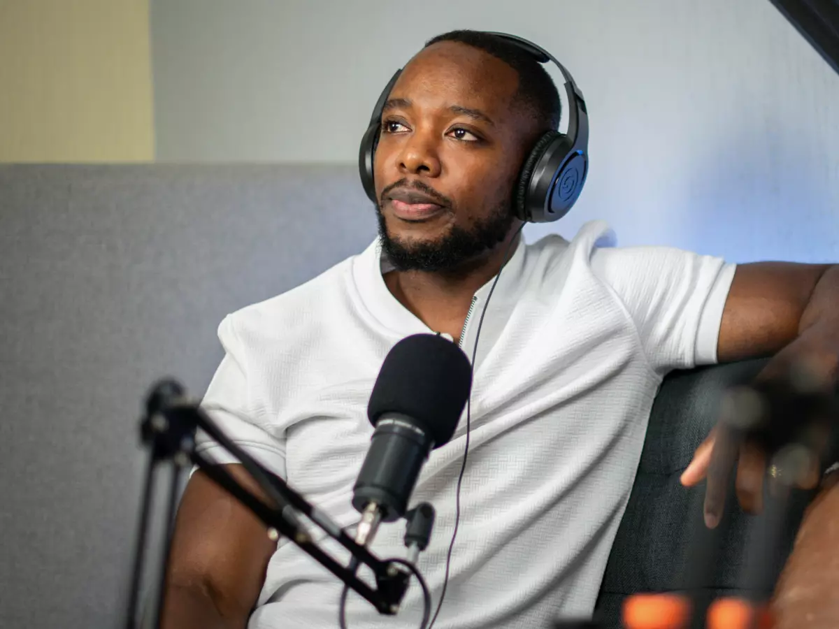 A man in a white t-shirt is recording a podcast in a home studio, with a microphone, headphones, and a laptop in front of him. He is sitting on a couch in front of a sound-dampening panel.